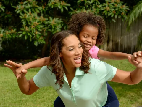 A mother carries her daughter on her back while playing outside.