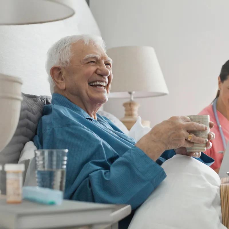 An elderly man seating on his bed in company of a nurse