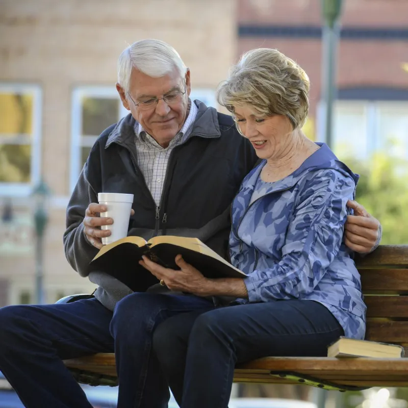 Elderly couple sitting on a park bench reading the bible.