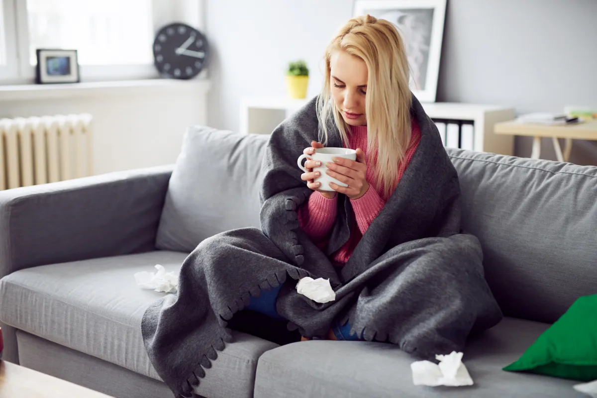 A woman drinking tea under a blanket at home.